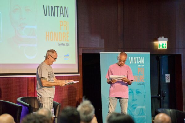 Deux intervenants lisent des textes sur scène lors des Vintan du Prix Honoré. L'un porte un t-shirt gris avec des inscriptions et un pantalon à carreaux, tandis que l'autre est vêtu d'un t-shirt rose et d'un jean. En arrière-plan, un écran affiche le texte 'Vintan Pri Honoré LanKrÉol 2024' sur un fond bleu avec un portrait en filigrane.
