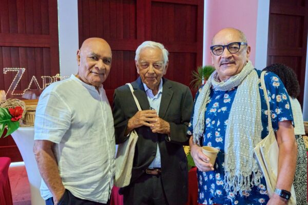 Trois hommes posent pour une photo lors des Vintan du Prix Honoré. L'un porte une chemise blanche, un autre un costume sombre avec un sac en toile sur l'épaule, et le troisième est vêtu d'une chemise à fleurs bleue avec une écharpe beige et tient un gobelet en carton. En arrière-plan, une table décorée de plantes et des lettres lumineuses formant le mot 'Zapero' sont visibles."