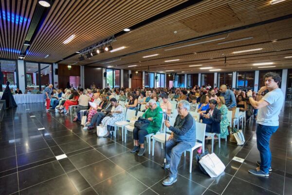 Vue de la salle lors des Vintan du Prix Honoré avec un public nombreux assis sur des chaises blanches, écoutant attentivement. Certains prennent des notes ou des photos avec leur téléphone. Un homme en t-shirt blanc se tient debout sur le côté avec son téléphone pour filmer l'événement. Le plafond est décoré de lames en bois et des spots lumineux éclairent la salle