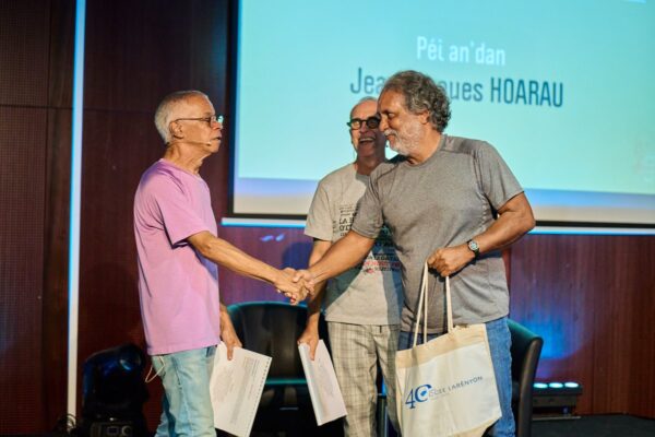 Deux hommes échangent une poignée de main sur scène lors de la remise des prix Vintan Pri Honoré 2024. L'un porte un t-shirt violet et tient un document, l'autre un t-shirt gris et un sac aux couleurs du CCEE.