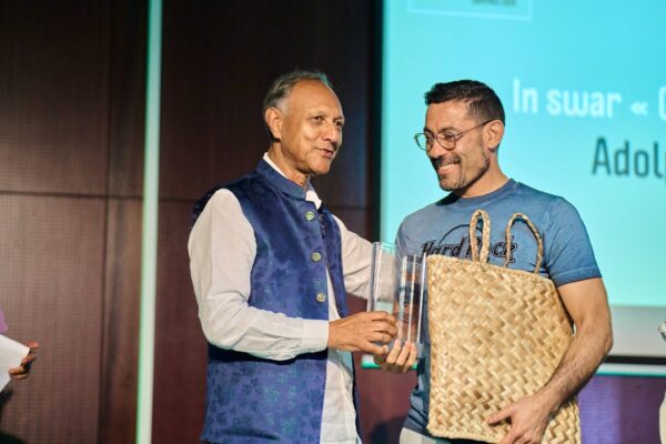 Un homme portant un gilet bleu remet un trophée Vintan Pri Honoré 2024 à un lauréat en t-shirt bleu, qui tient également un panier en vacoa. Les deux hommes échangent un sourire chaleureux sur scène.