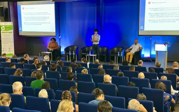 Vue générale de la salle de conférence avec le public attentif aux intervenants sur scène, écrans projetant des diapositives