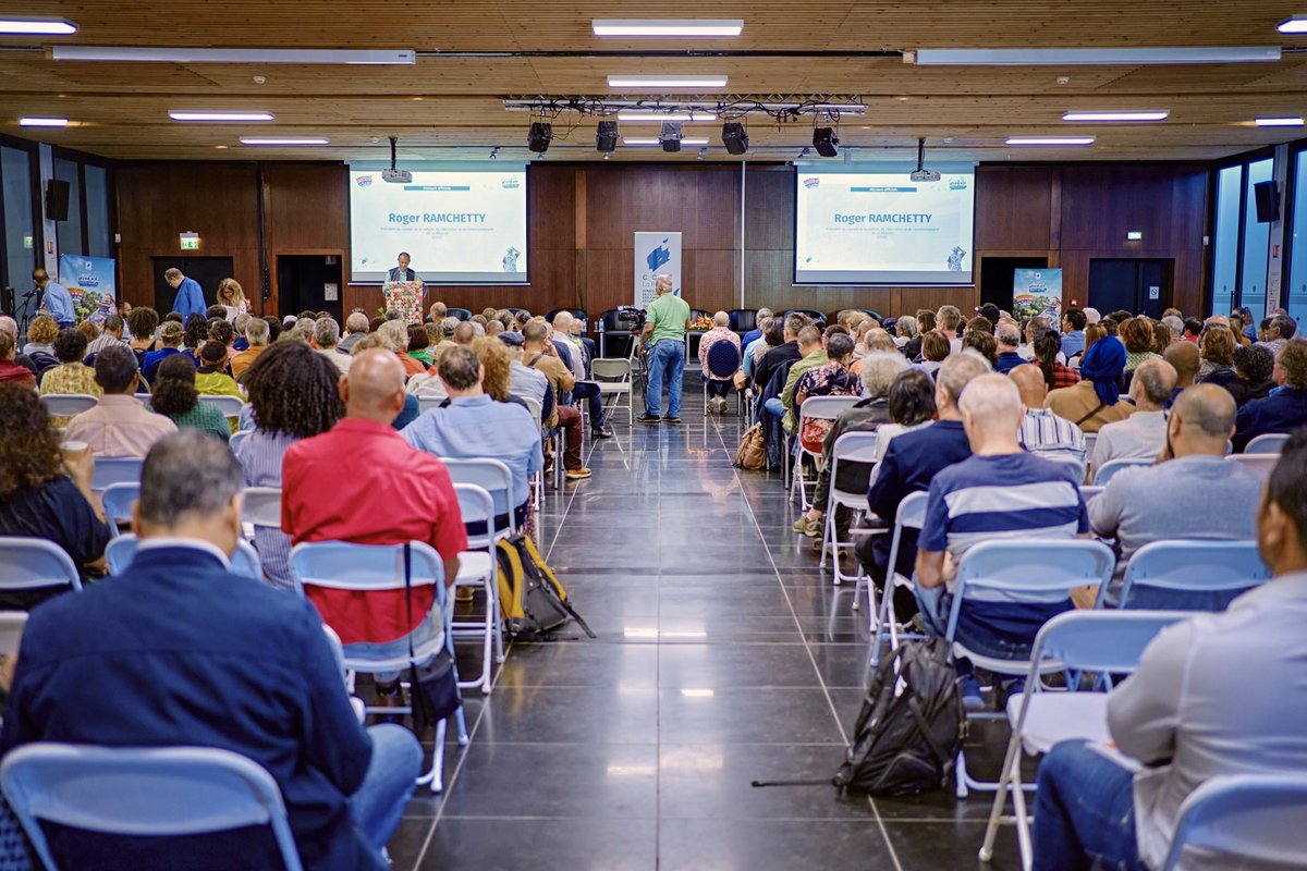 Salle comble lors d'une conférence, vue arrière des participants face à un intervenant sur scène.