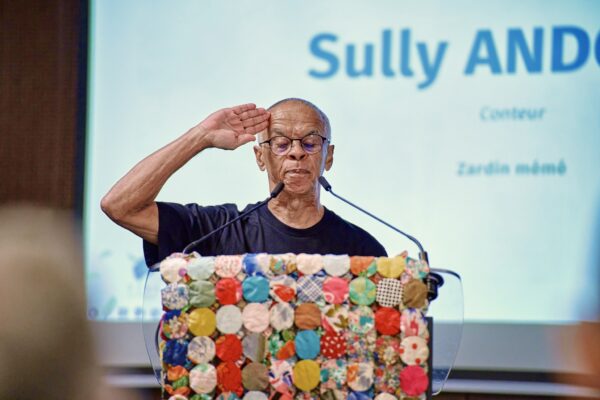 Sully Andoche saluant la salle d’un geste militaire, debout au pupitre, avec son nom "Sully Andoche" projeté à l’écran.