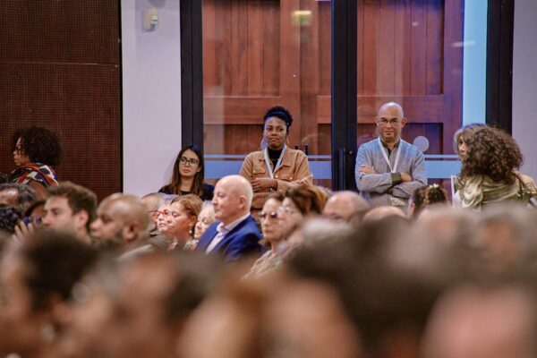 Trois personnes debout au fond d’une salle de conférence, observant l’événement en cours.