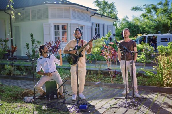 Trio de musiciens en extérieur, avec un guitariste chantant, un percussionniste et un homme au micro.