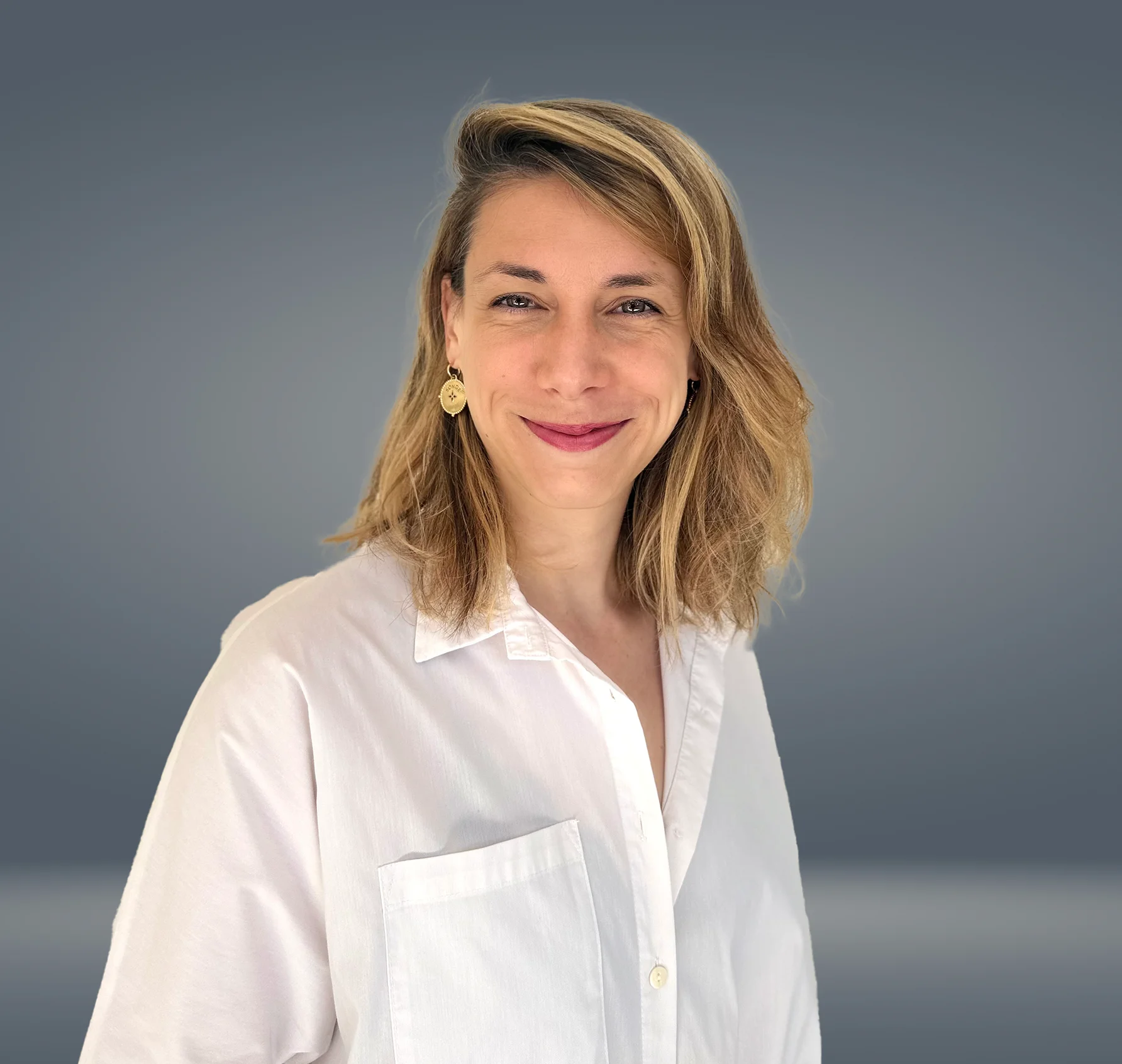 Portrait de Cyrielle Jacheet, une femme souriante aux cheveux blonds mi-longs, portant une chemise blanche et des boucles d’oreilles dorées, sur un fond uni gris dégradé.
