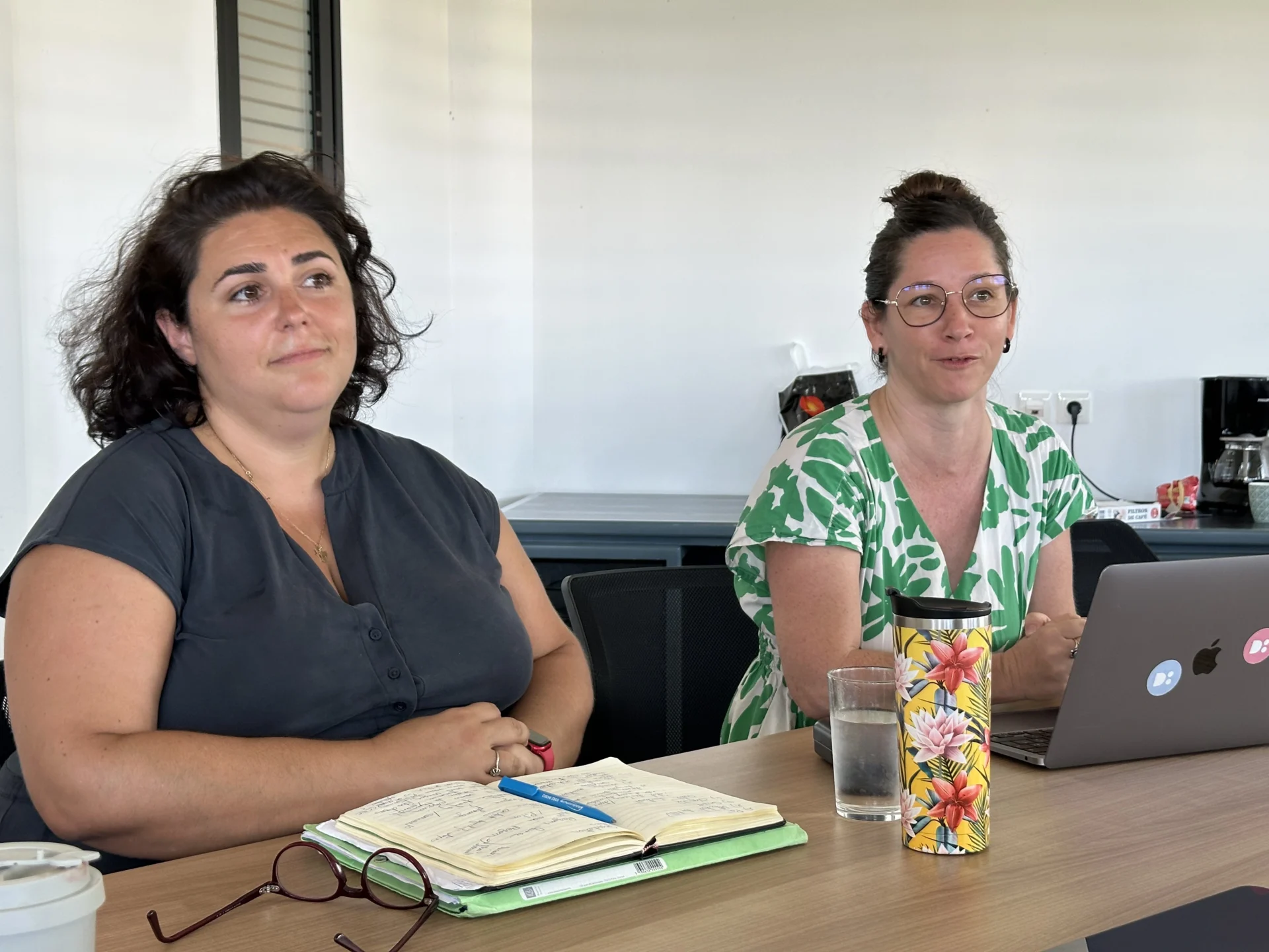 Deux femmes participent à une réunion de travail autour d'une table avec un carnet de notes ouvert.