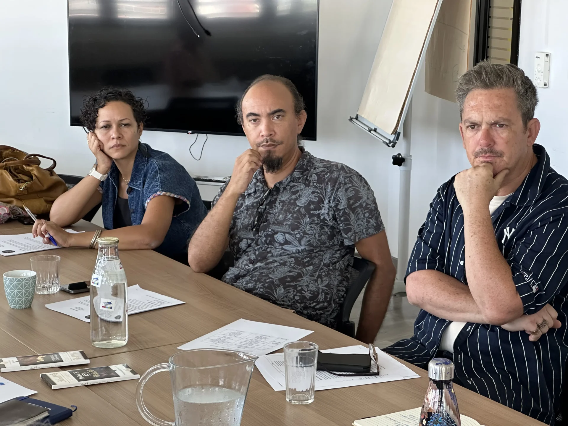 Trois personnes, une femme et deux hommes, sont assis côte à côte lors d'une réunion. Ils affichent une expression attentive et concentrée. Des documents de travail et des bouteilles d'eau sont posés devant eux sur la table. Un écran de télévision éteint et un tableau de conférence sont visibles en arrière-plan.