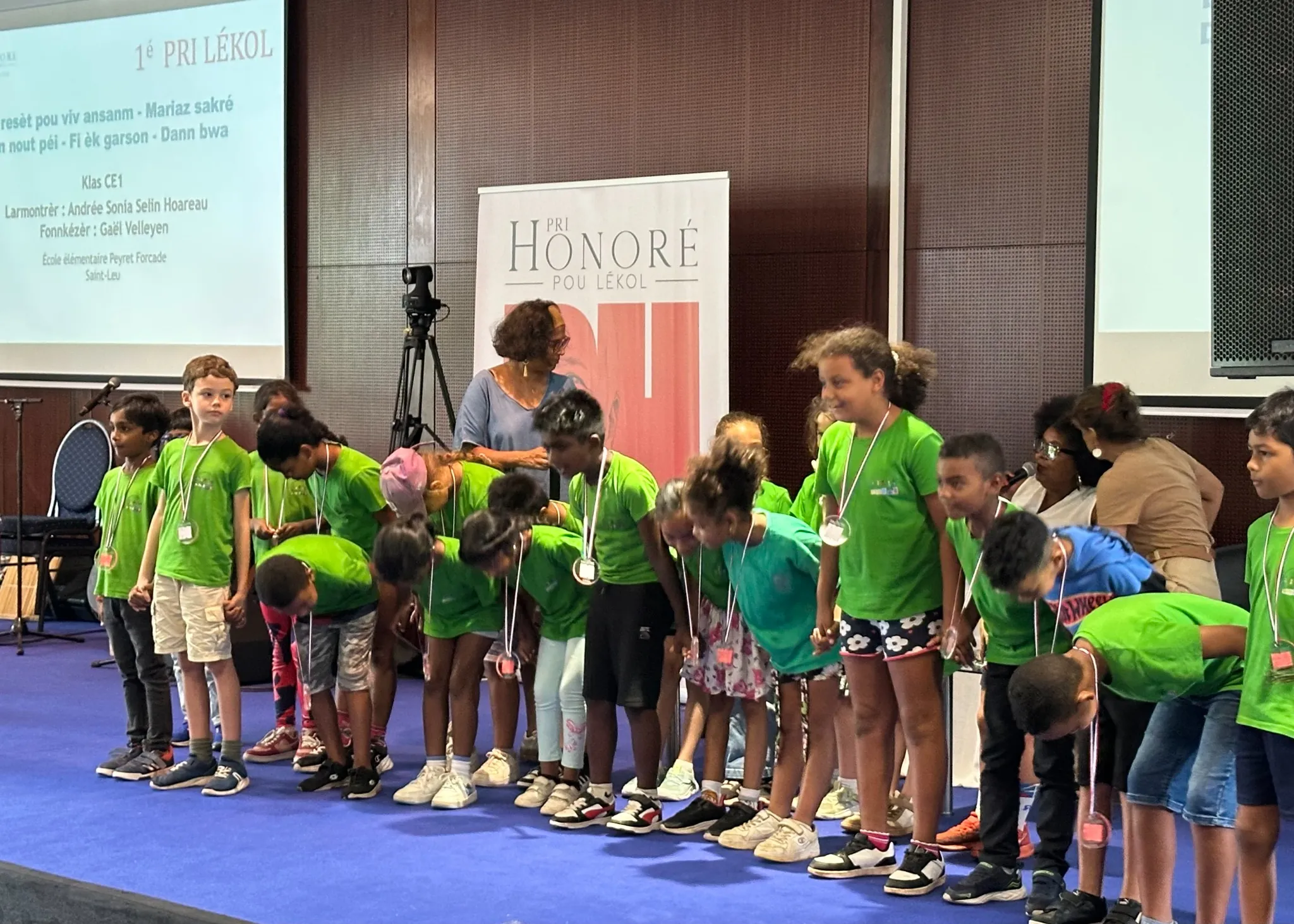 Les élèves lauréats du 1er Prix Lékol, en t-shirts verts avec leurs médailles, sur scène au MoCA lors de la cérémonie du Pri Honoré pou lékol 2026, devant le roll-up officiel de l'événement